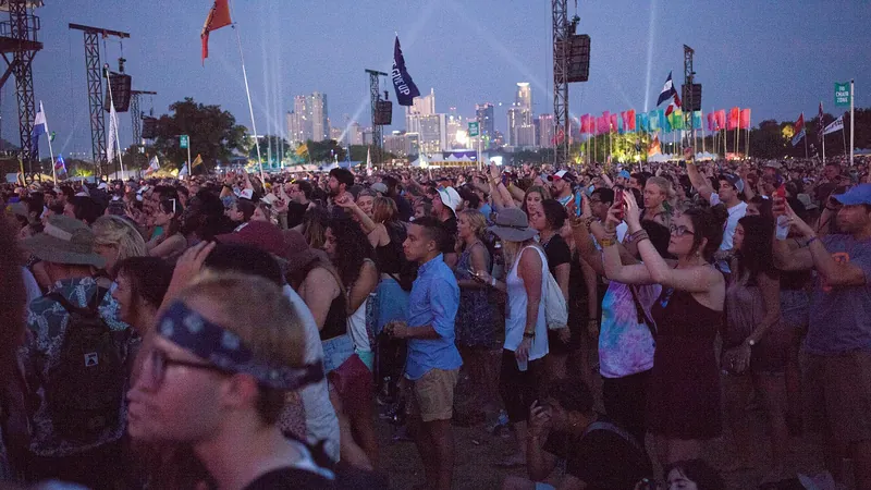 Multitud en un festival al aire libre al atardecer, con banderas y focos sobre el horizonte