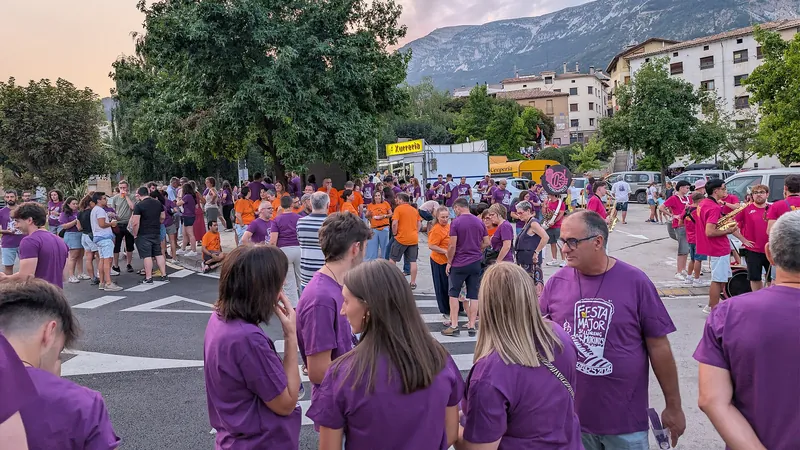 Peñas con camisetas moradas reunidas en la plaza de un pueblo de montaña durante las fiestas mayores