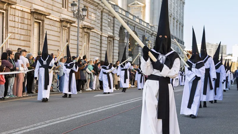 Fila de nazarenos con túnica blanca y capirote negro desfilando junto al Palacio Real de Madrid